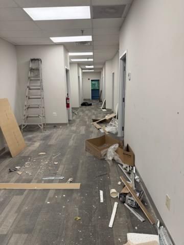 Messy office hallway under renovation with ladder, scattered debris, and ceiling lights on.