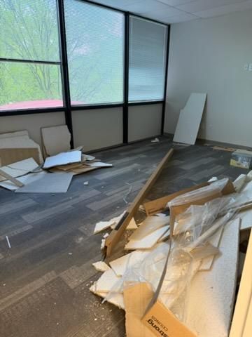 Damaged office with scattered broken furniture and debris on the floor near large windows.