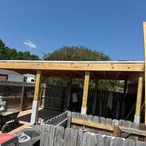 Backyard patio with wooden pergola, fence, and trees under a clear blue sky