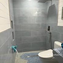 Small bathroom with gray tiled walls, walk-in shower, toilet, and mop on the floor.