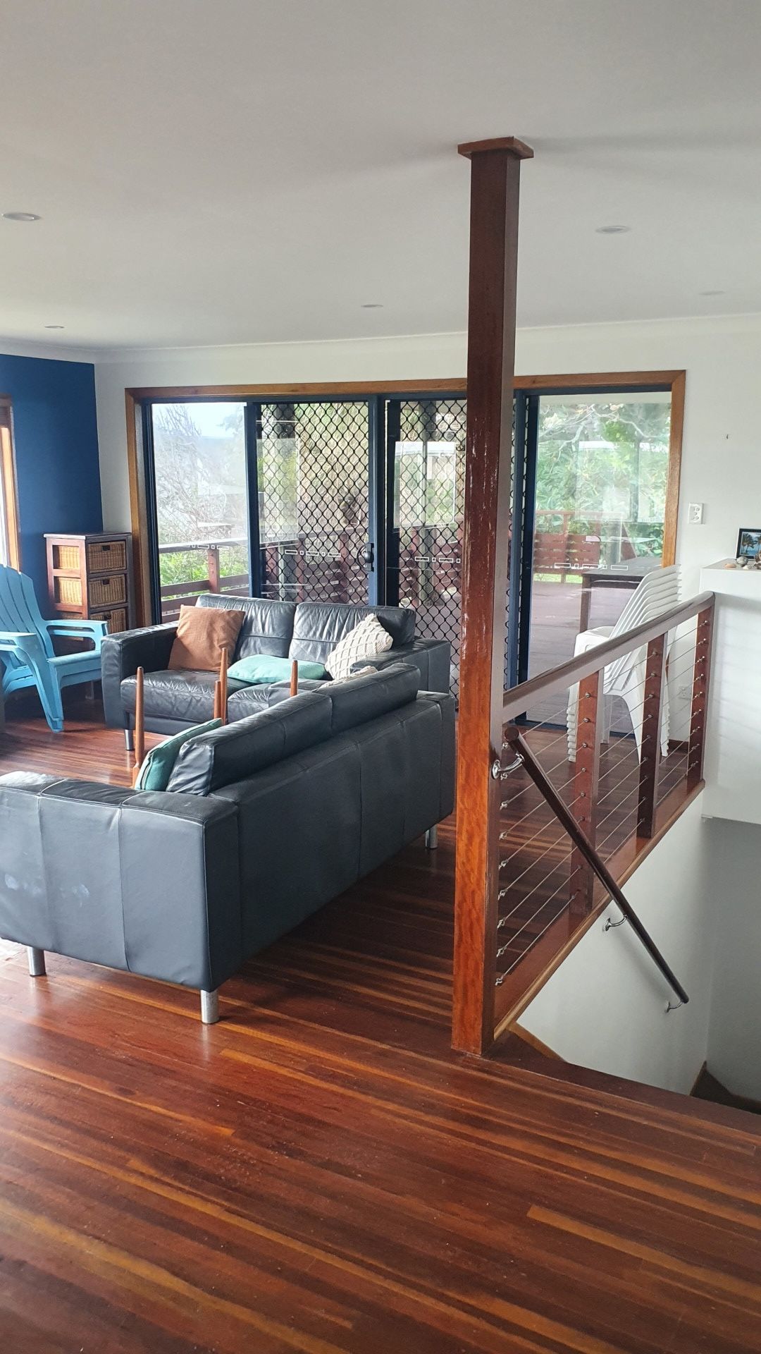 Living room with hardwood floors, black leather sofa, deck access, wooden staircase, and a wooden support column.— Keppel Coatings in Pacific Heights, QLD