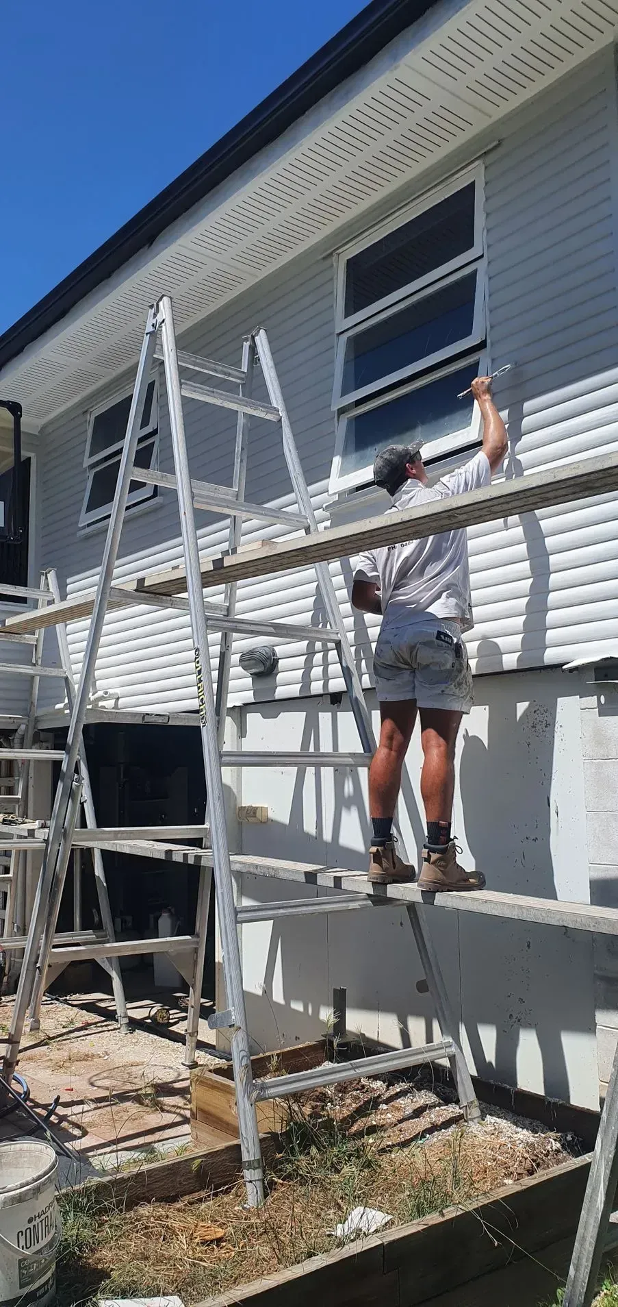Person Painting a House. They Are on a Scaffold Next to a Window — Keppel Coatings in Pacific Heights, QLD