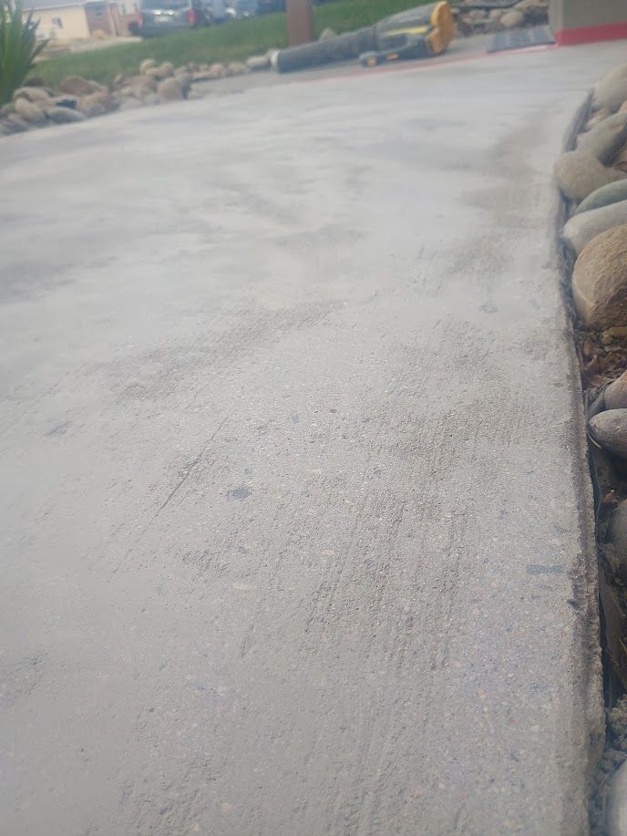 Concrete driveway beside a landscaped edge with rocks and curb, leading toward a building and parked equipment.