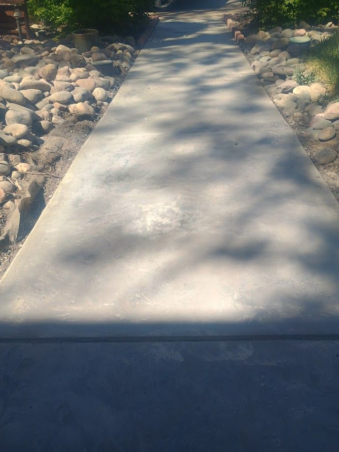 Concrete walkway bordered by rocks and greenery, with sunlight and shadows across the path