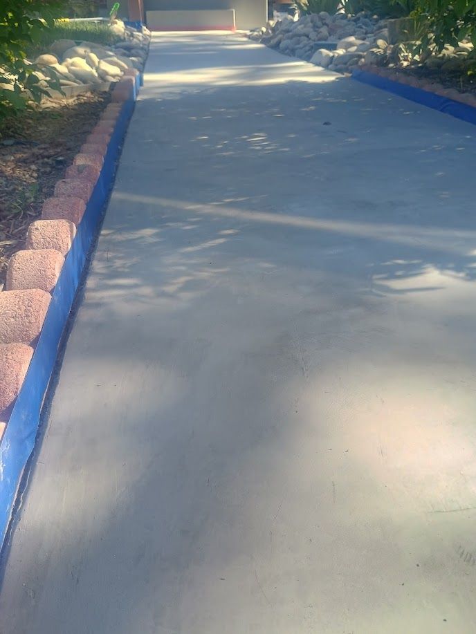Concrete walkway bordered by blue curbs and red pavers, shaded by trees in a garden path.