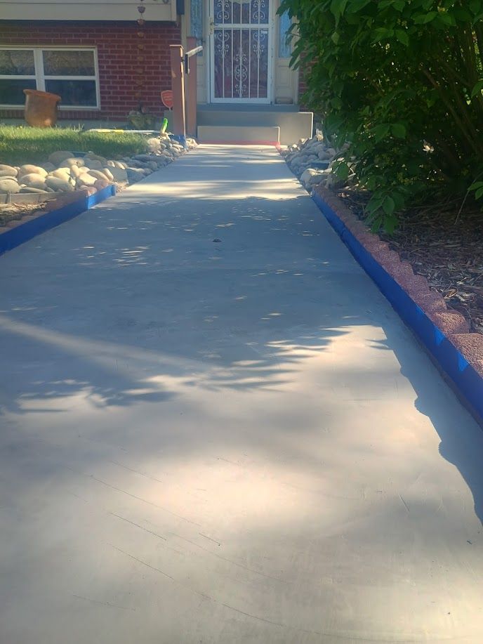 Concrete walkway leading to a house entrance, bordered by blue curbs and shrubs in sunlight.