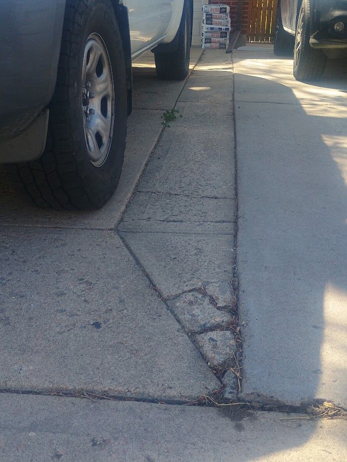 Cracked sidewalk beside parked cars, with a loose triangular concrete slab.
