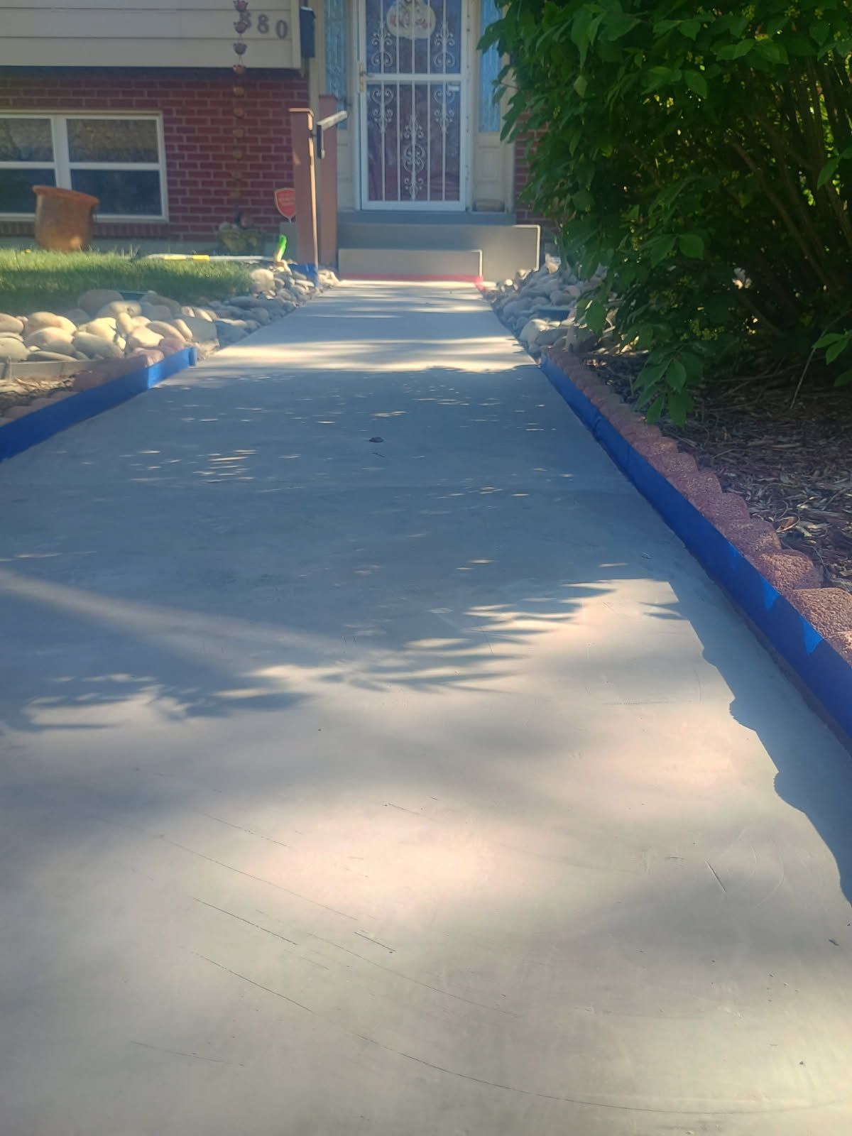 Concrete walkway leading to a front porch, bordered by rocks and blue-painted edges in bright sunlight