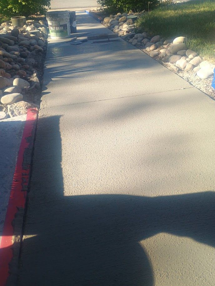 Sunlit concrete walkway bordered by rocks and shrubs, with a red curb line on the left