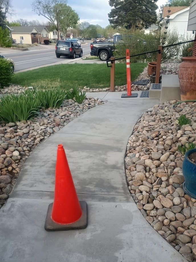Concrete walkway under construction with orange cones and red caution tape in a landscaped front yard.