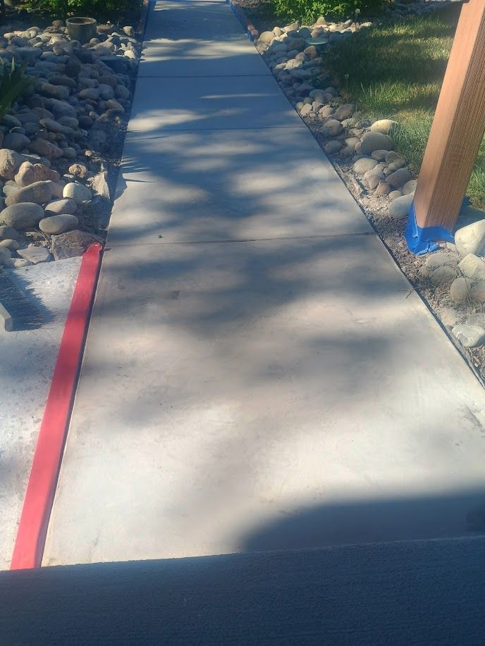 Concrete sidewalk path with a red curb line, rocks on both sides, and dappled sunlight.