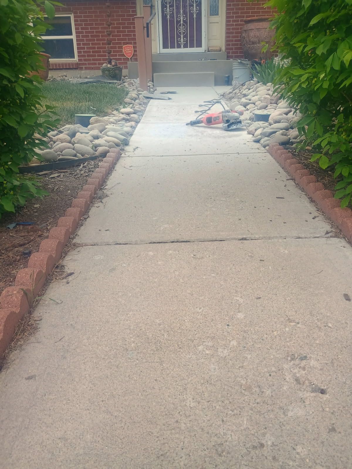 Concrete walkway leading to a brick house entrance, lined with rocks and shrubs