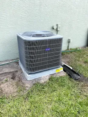 A grey Carrier air conditioning unit sits on a concrete pad against a light green stucco wall in a grassy yard.