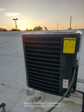 A dark grey air conditioning unit sits on a flat roof during a sunset, featuring a prominent yellow EnergyGuide label.