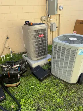 Two outdoor HVAC units sitting on a concrete pad next to a beige exterior wall, with maintenance equipment on the grass.