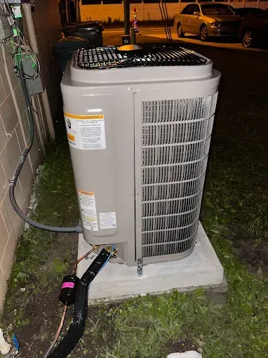 An outdoor central air conditioning unit sits on a concrete pad next to a brick wall at night, with a car in the background.