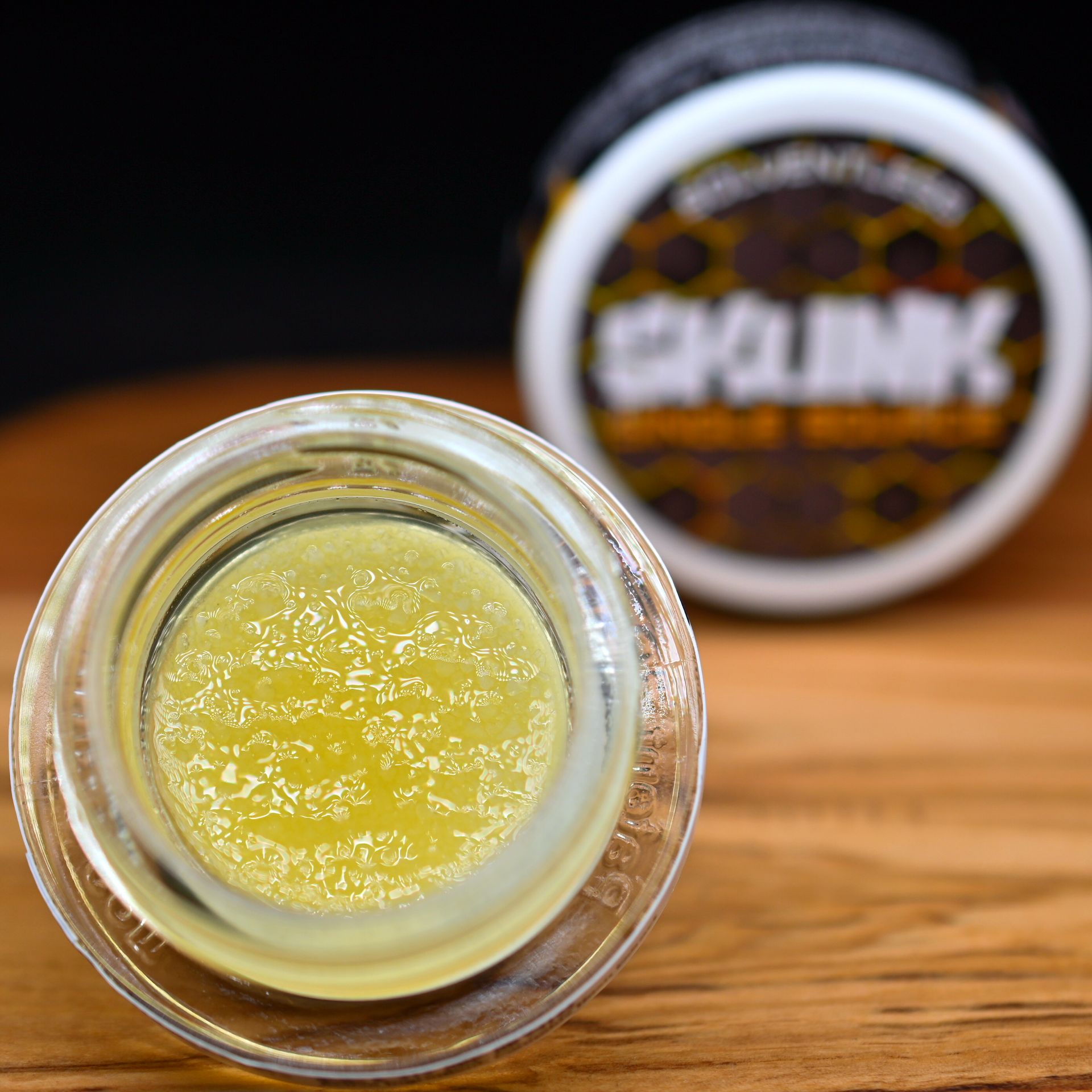 Open glass jar of yellow balm on a wooden surface, with blurred labeled container in the background