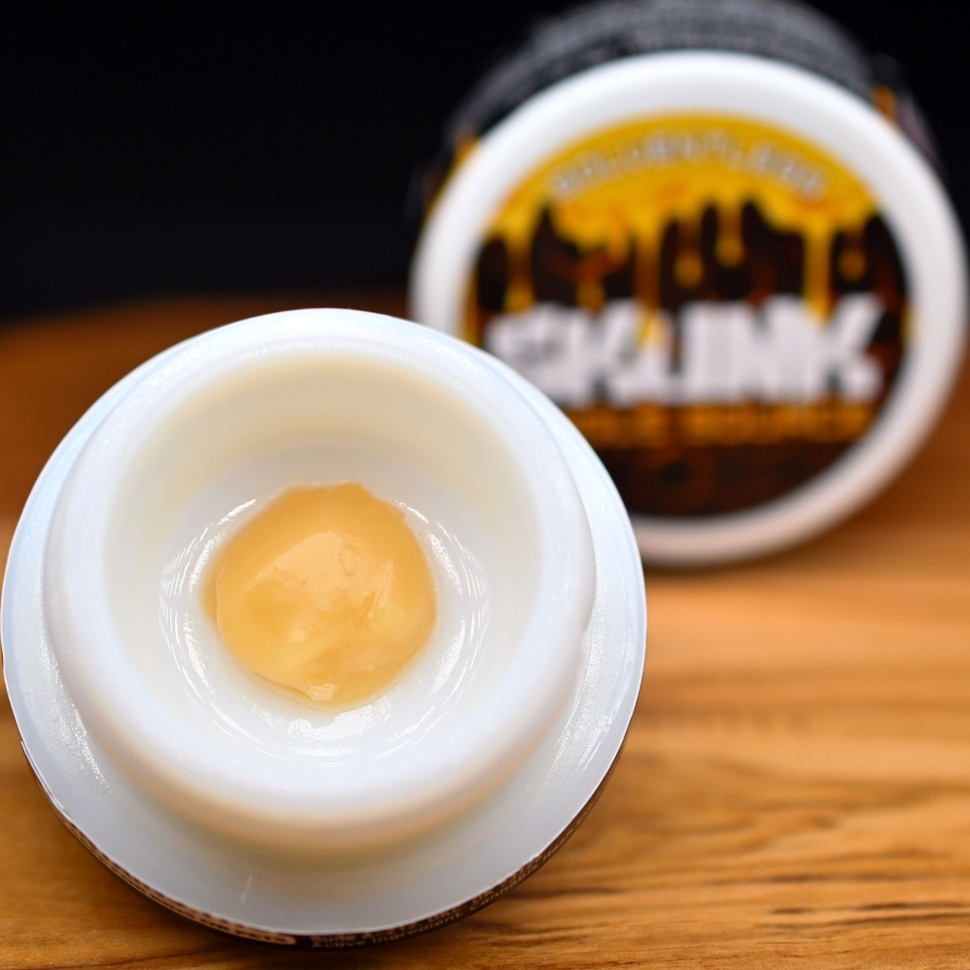 Open jar of pale yellow balm on a wooden table, with labeled lid blurred in the background