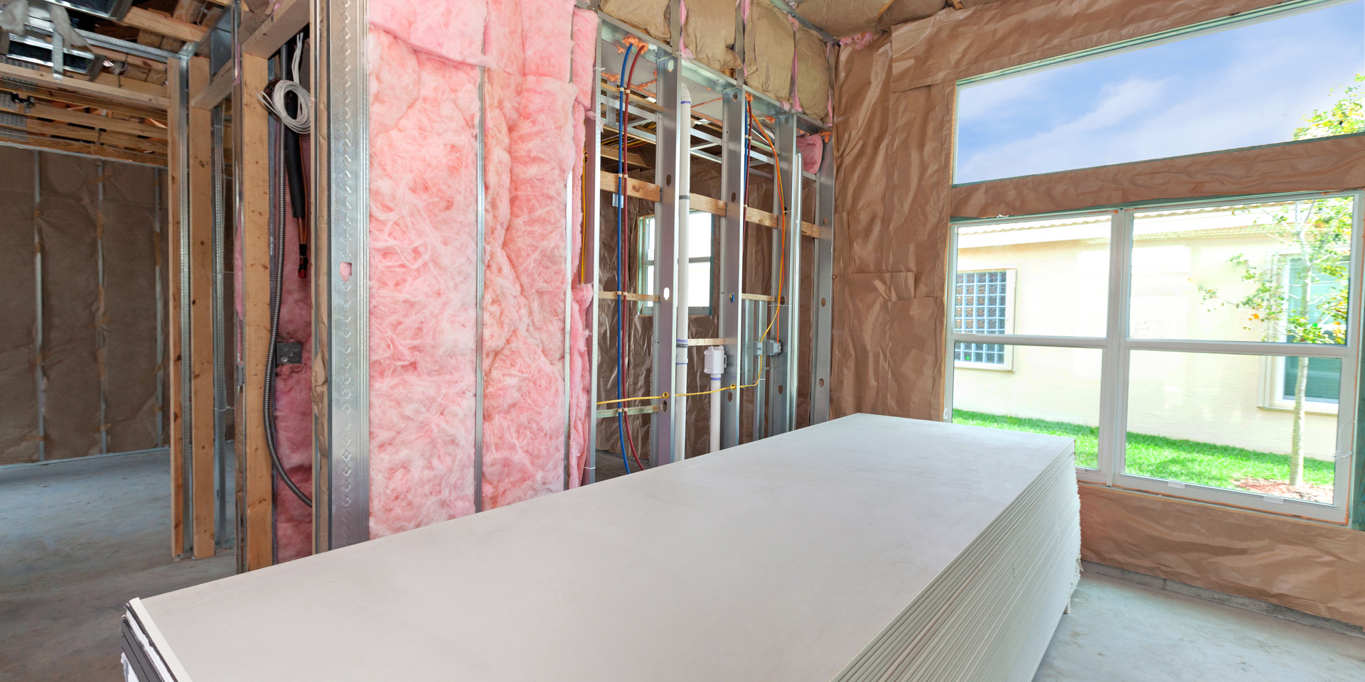 Interior of a building under construction, showing metal framing, insulation, and drywall. A window offers a view outside.