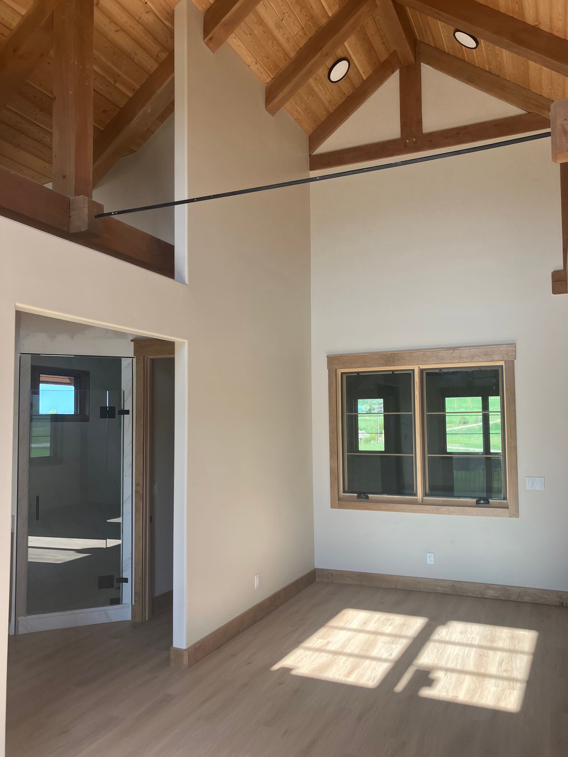 Interior view of a room with light wood floors, beige walls, and a wood-beamed ceiling. Sunlight streams through a window.