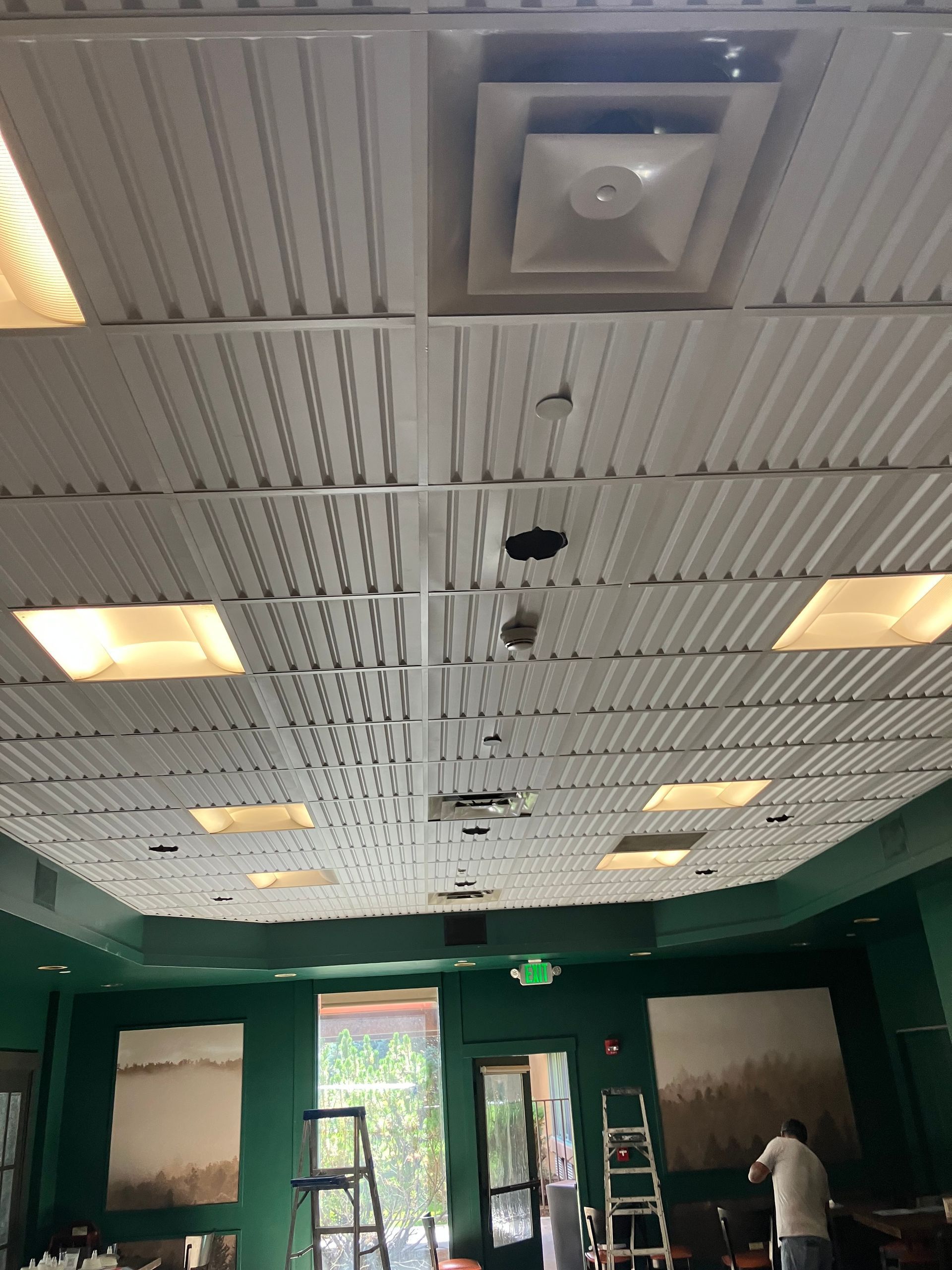 White corrugated metal ceiling with vents and lighting fixtures. Green walls, and a person on a ladder in a room.
