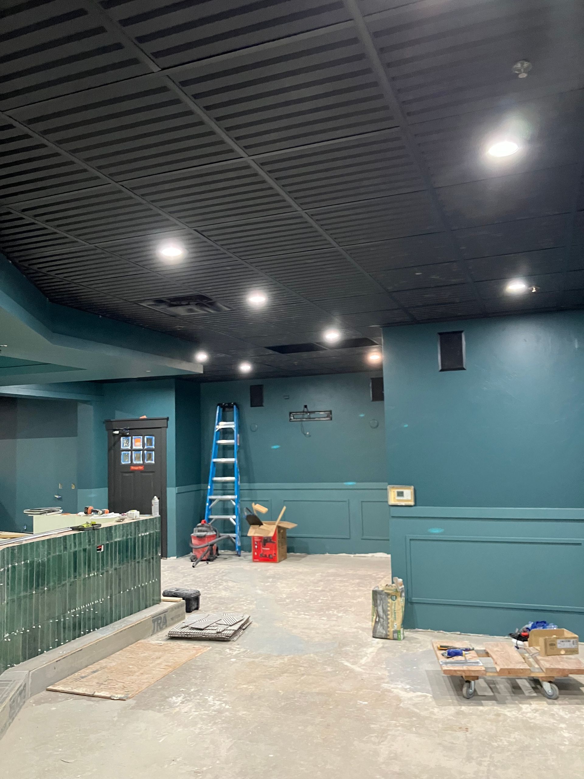 Interior of a teal room under construction; black ceiling, spotlights, a ladder, and tools are present.