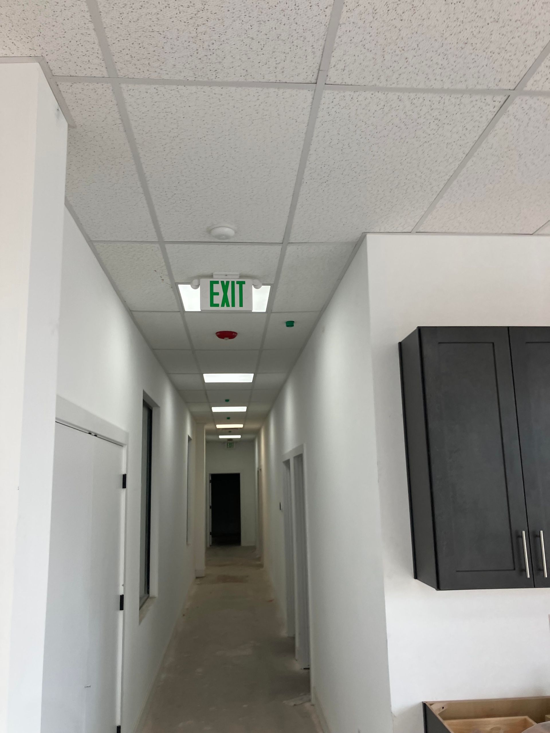 Long hallway with white walls, ceiling tiles, and an exit sign.