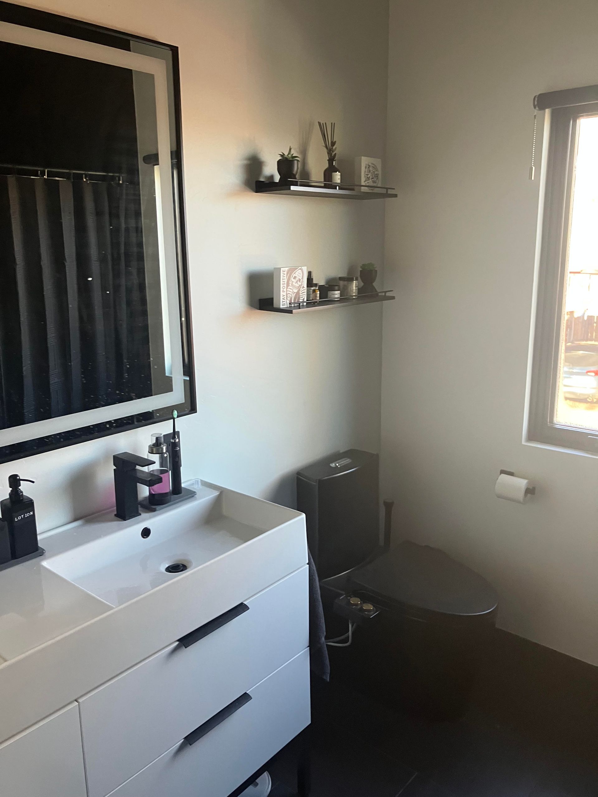 Modern bathroom with white vanity, black toilet, window, and shelves with decorative items.