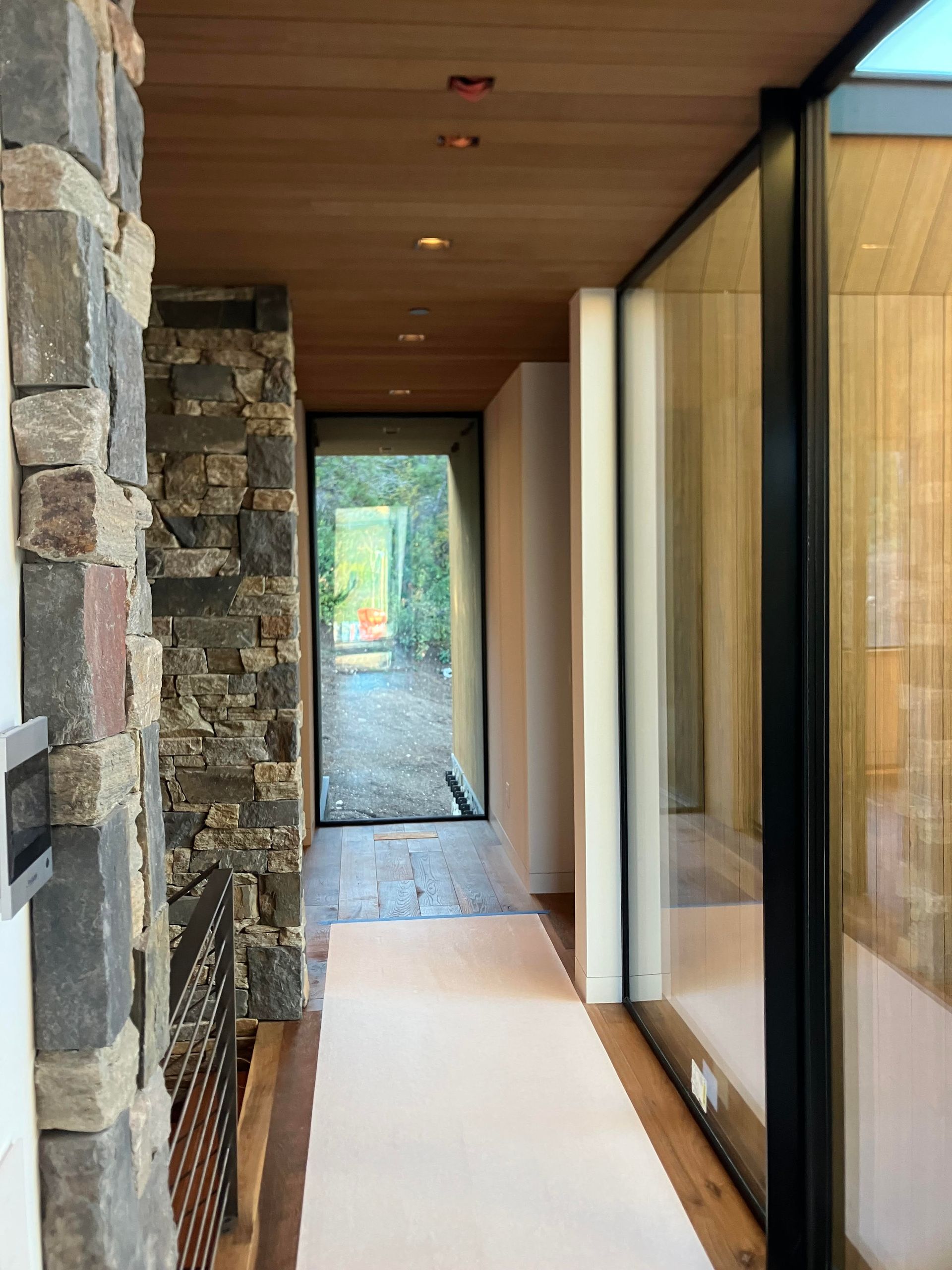 Stone-walled hallway with glass wall, leading to an outdoor view. Wooden ceiling and floor.