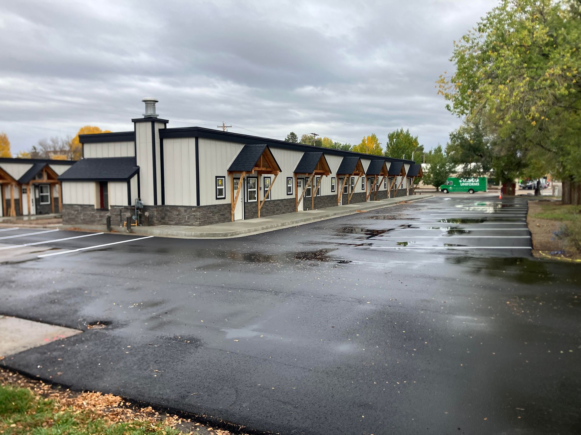 Motel building with black roofs, white siding, and stone accents. Wet asphalt parking lot. Overcast sky.
