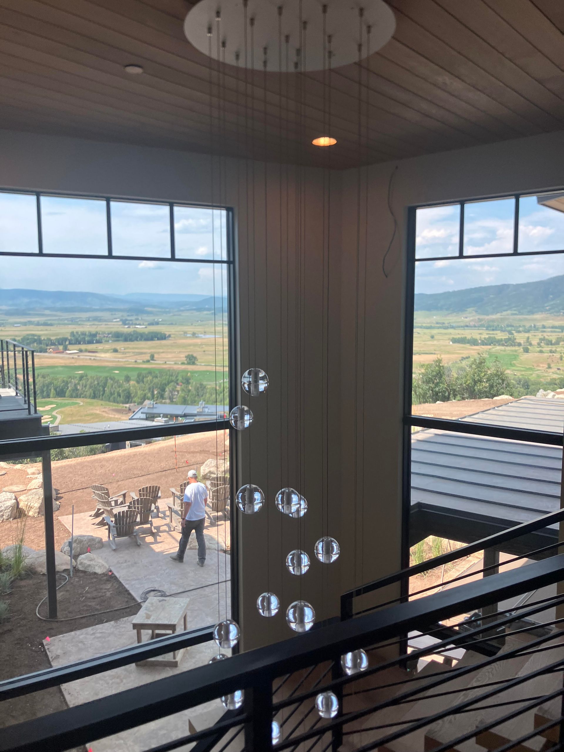 Modern interior with large windows overlooking a valley. A chandelier hangs; a person walks outside.