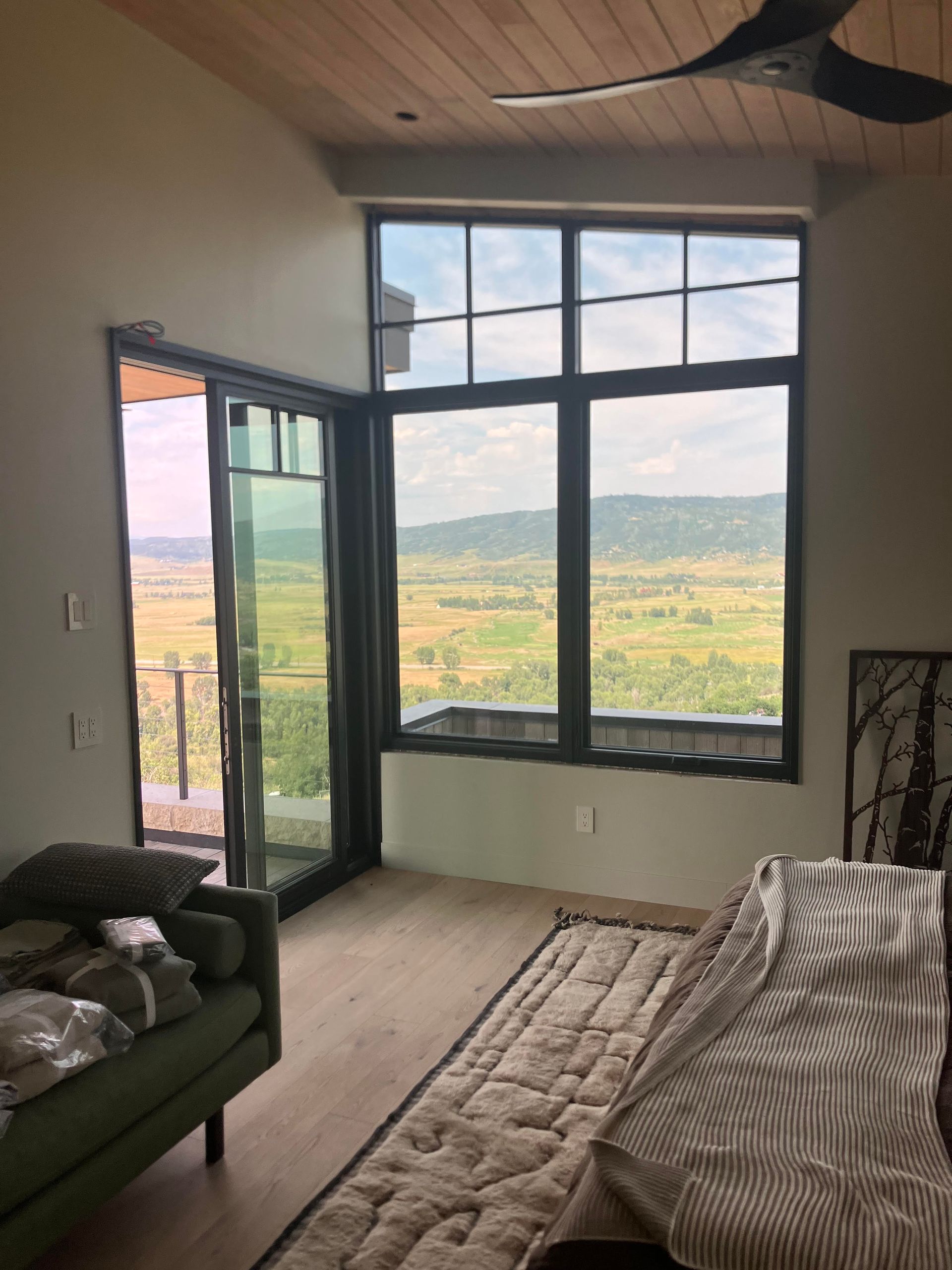 Room with a large window and sliding glass door overlooking a valley. The room has light-colored wood flooring, and a green couch.