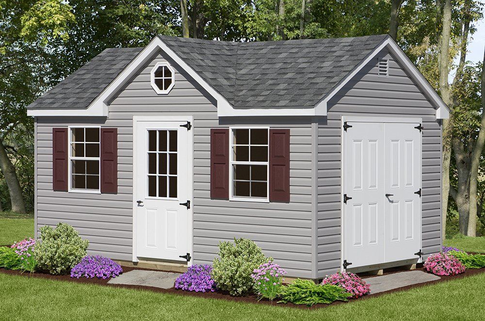 Gray shed with dark roof, white doors, and burgundy shutters on a grassy lawn with flowers.