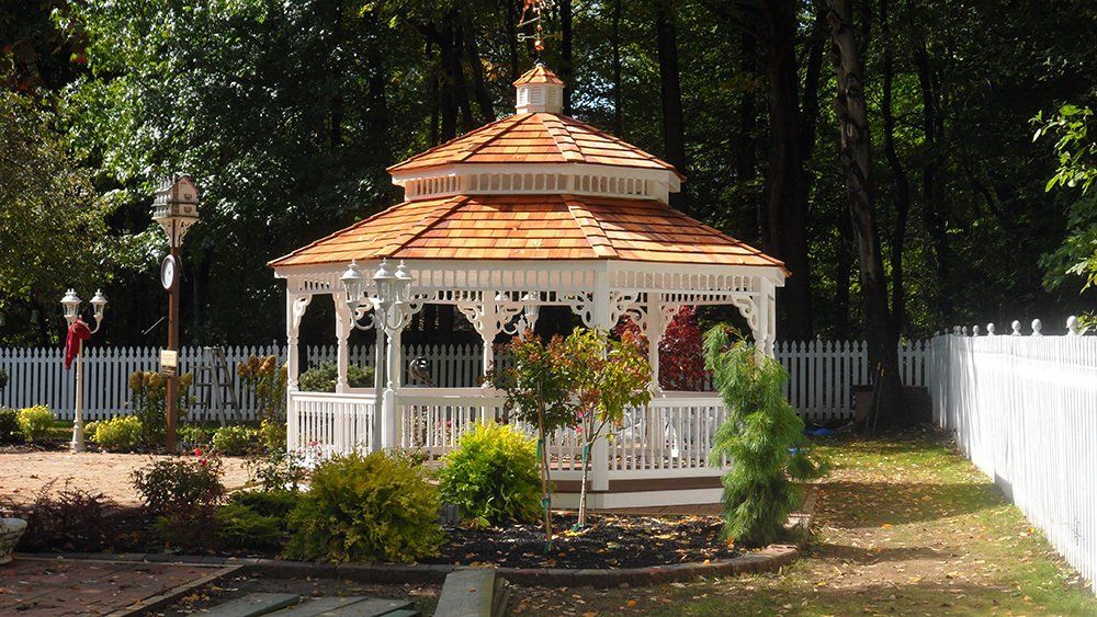 White gazebo with brown roof in a garden, surrounded by trees and a white picket fence.