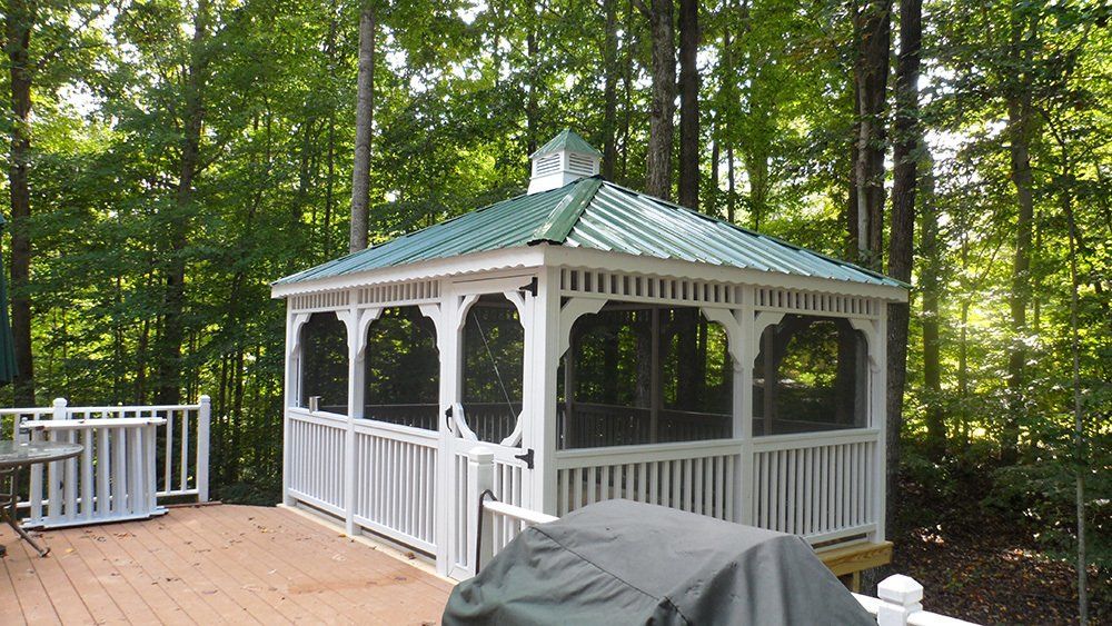 White gazebo with green roof, screened windows, on a wooden deck surrounded by trees.