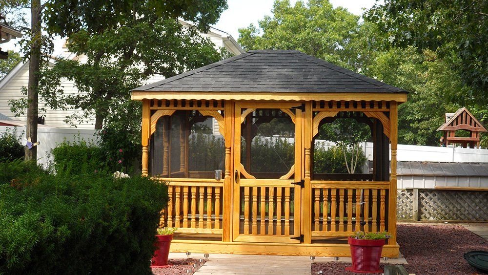 Wooden gazebo with black roof in a backyard, surrounded by trees and shrubbery.