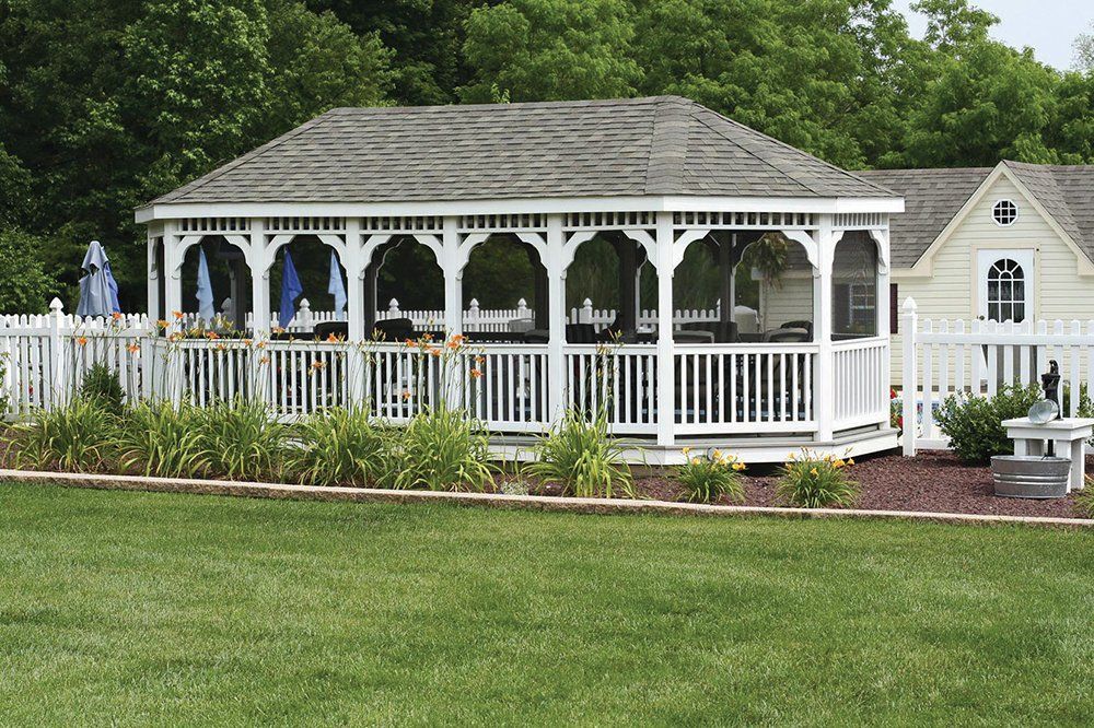 White gazebo with screened openings and a gray roof, surrounded by a white picket fence, green lawn, and trees.