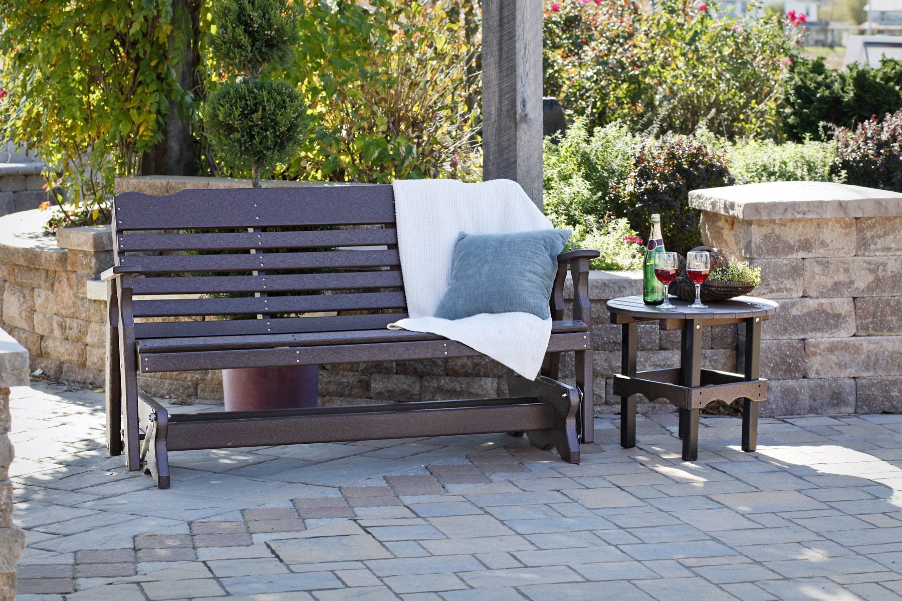 Outdoor brown gliding bench with a side table, blanket, and pillow, on a patio.