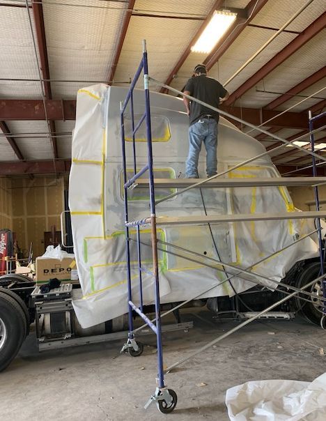 Person on scaffolding prepping a truck cab for paint in a workshop; masked and covered.