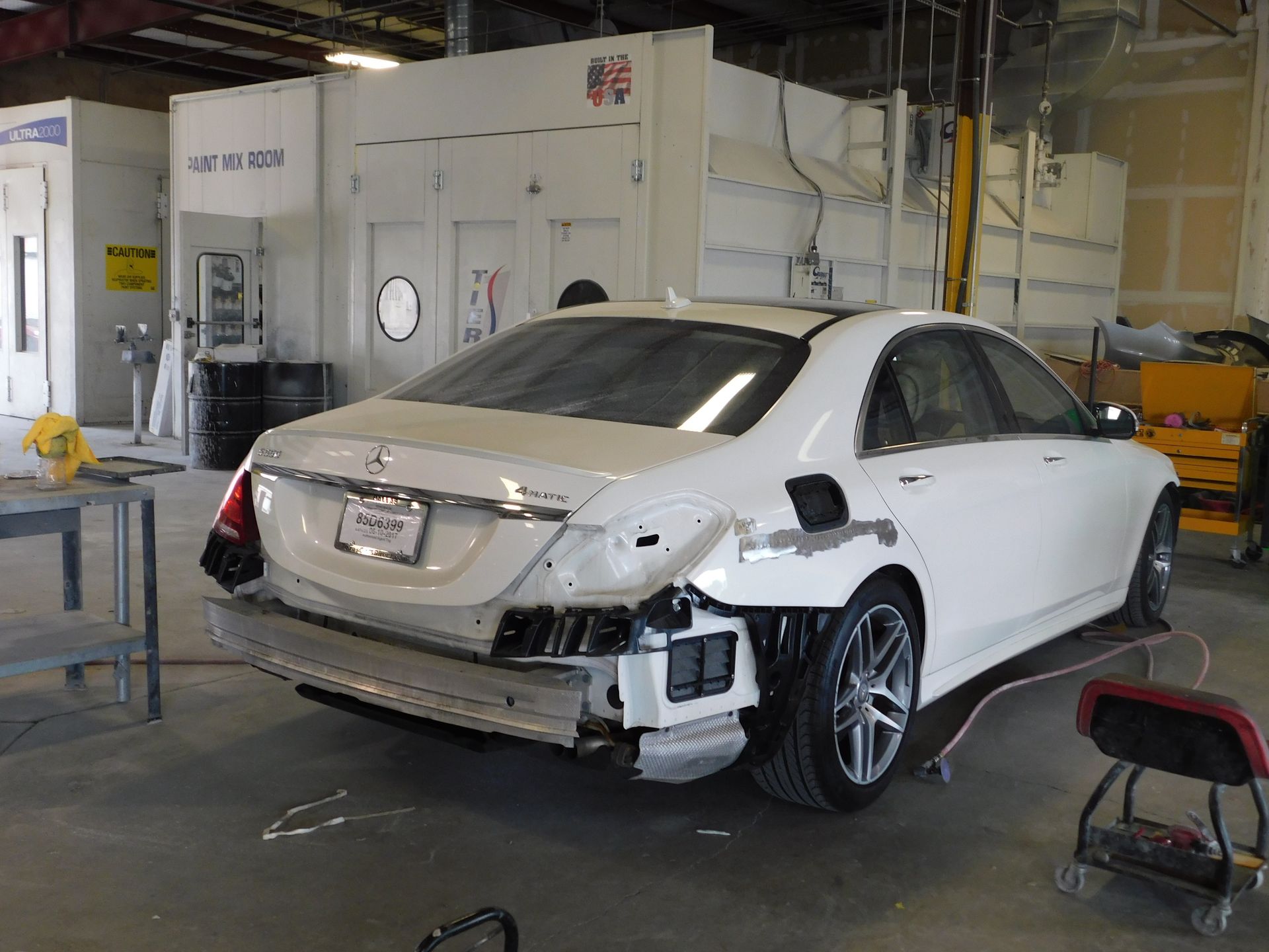 White Mercedes sedan in auto body shop, rear bumper removed.