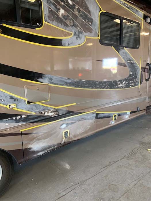 RV side undergoing repair, masked for paint, with areas of exposed bodywork and a brown and black design.