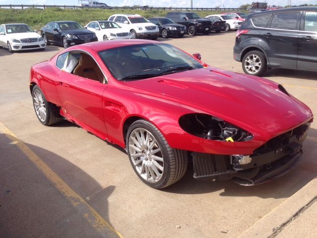 Damaged red Aston Martin car with missing front parts, parked in a lot.