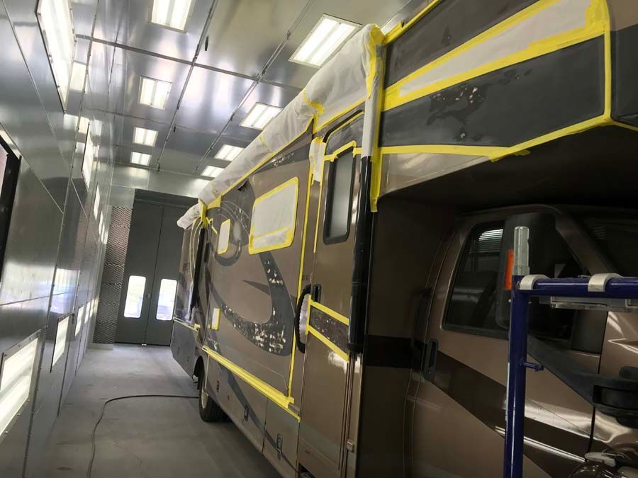 RV in a paint booth, taped for painting. Beige and brown with yellow tape, inside a bright white setting.