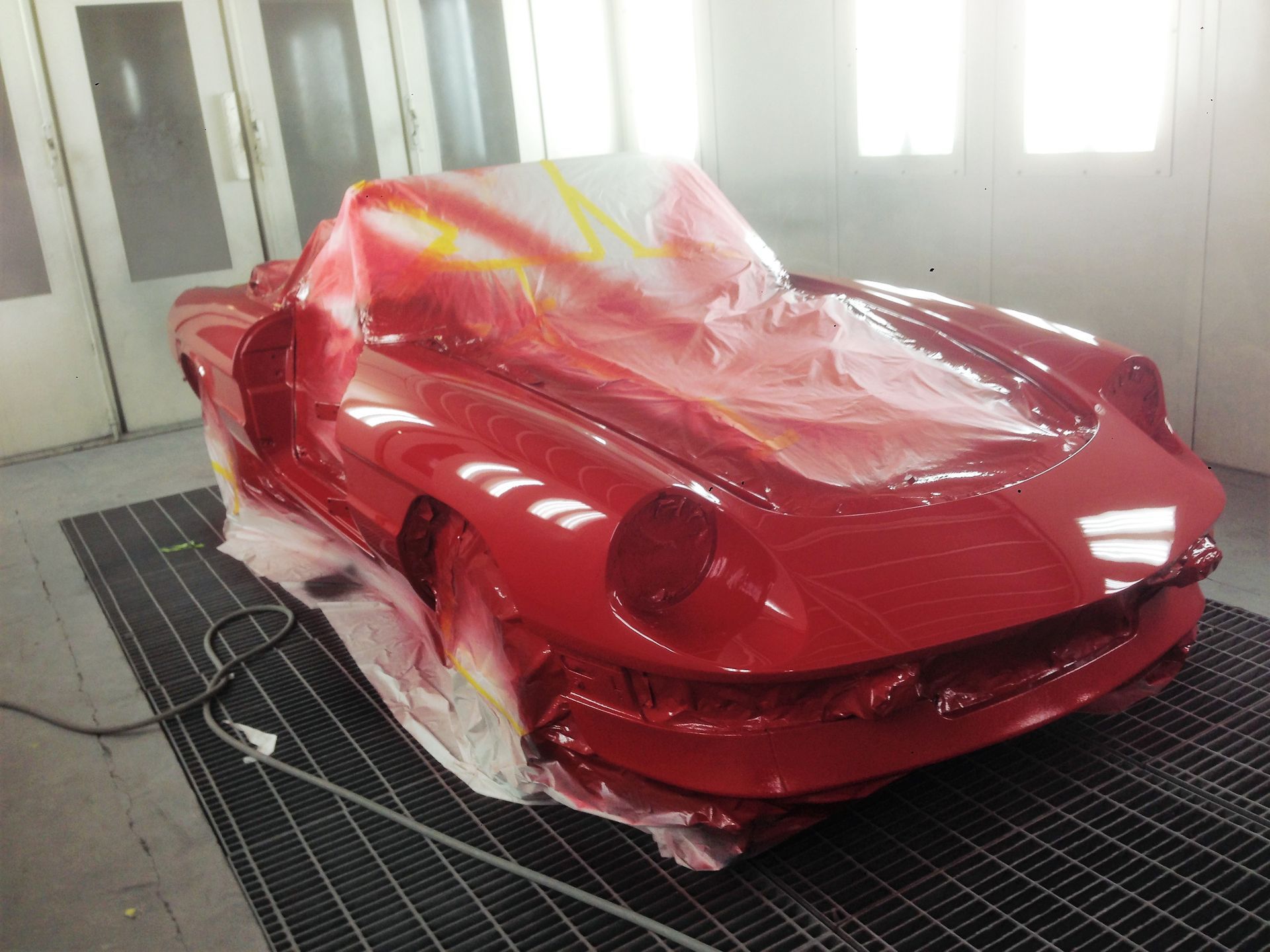 Red sports car being painted in a body shop, masked and covered in plastic.