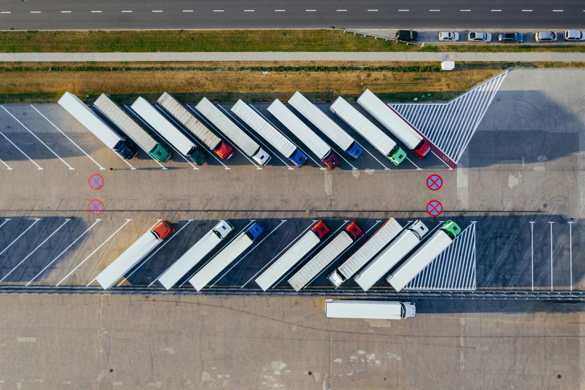 Overhead view of parked semi-trucks in rows at a truck stop. Trailers are white, cabs are various colors.