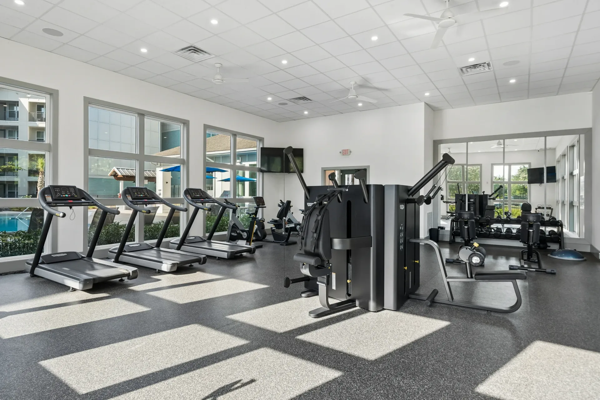 Modern apartment gym with treadmills, weight machines, and free weights, overlooking the pool area.