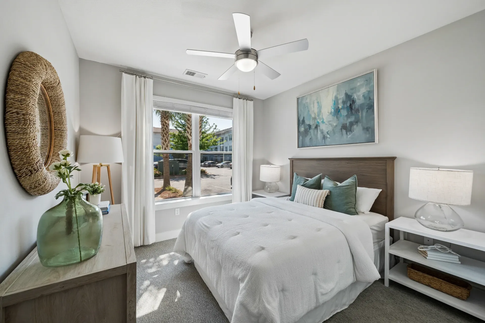 Bright bedroom with a queen-size bed, white linens, and teal accent pillows. Features a ceiling fan and a large window with a view of palm trees and buildings at Atlantic on the Boulevard in North Charleston, SC.