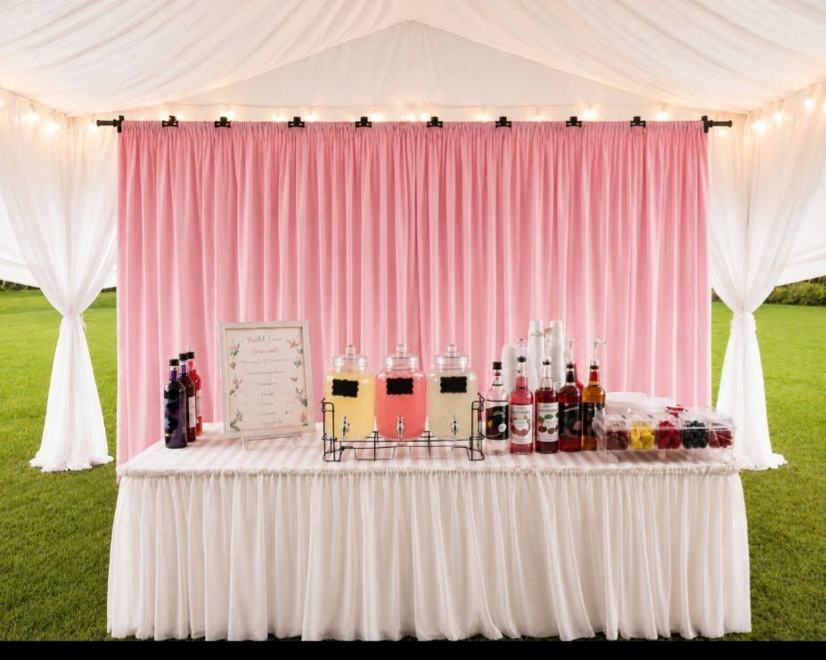 Outdoor drink bar with pink backdrop, white drapes, and assorted beverages on a decorated table