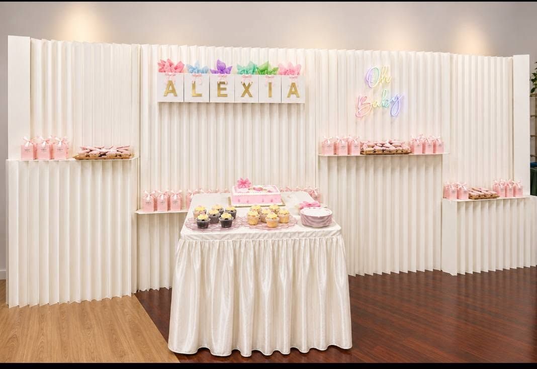 Birthday dessert table with white drapes, pink accents, and “ALEXIA” banner above cupcakes and cake