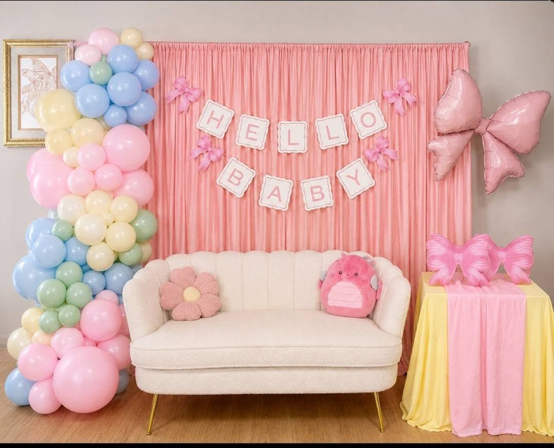 Pastel baby shower backdrop with balloons, pink fringe curtain, couch, and gift table